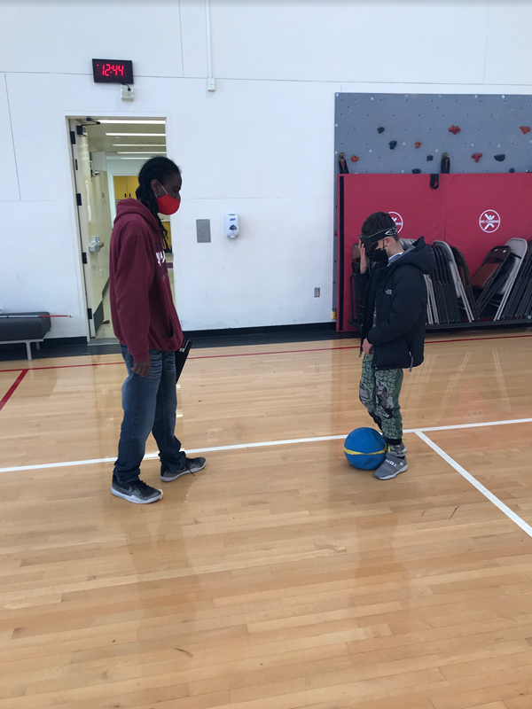 2 students are standing across from each other practicing passing a rubber ball with their feet. The student on the right is adjusting their blindfold. 